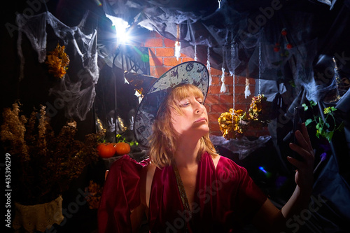 Blonde witch in red dress and black hat in Halloween decoration indoors with cell phone. Woman taking a selfie during holiday carnival