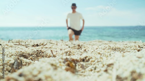Man is walking along the beach. Young man in a white T-shirt, black shorts and a black hat walks along the sand to the sea. Sunny summer day on the beach. Holidays in a warm country. Blue sea.