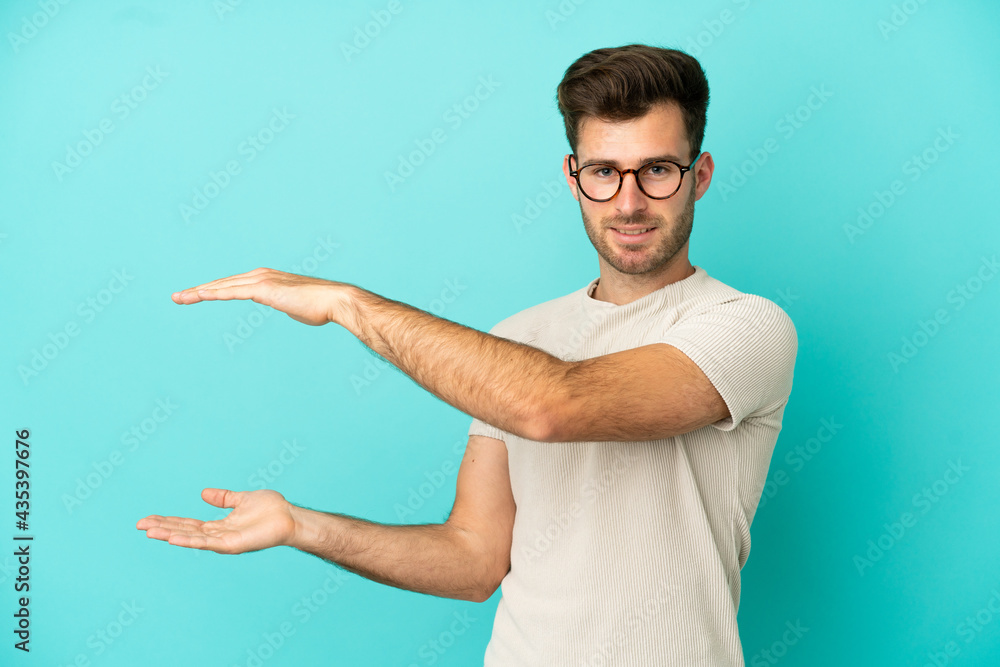 Young caucasian handsome man isolated on blue background holding copyspace to insert an ad Young caucasian handsome man isolated on blue background holding copyspace to insert an ad