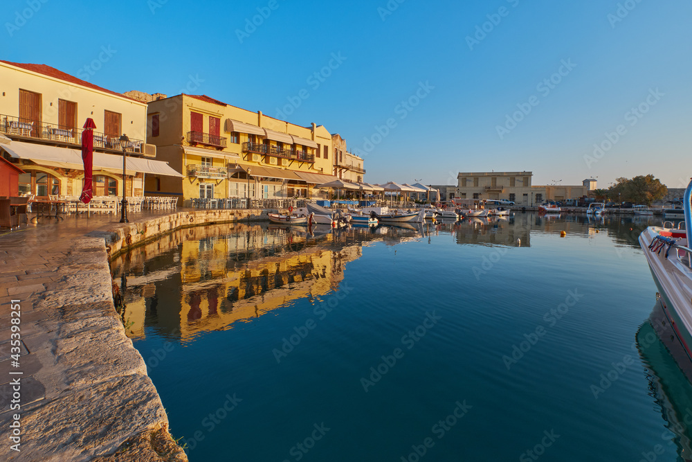 the quay of the historic harbour of Rethymno in the morning sun, boats in the port and still closed restaurants