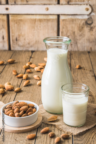 Glass and pitcher with vegan almond milk, almond kernels and whole almonds on an old rustic wooden table, vertical