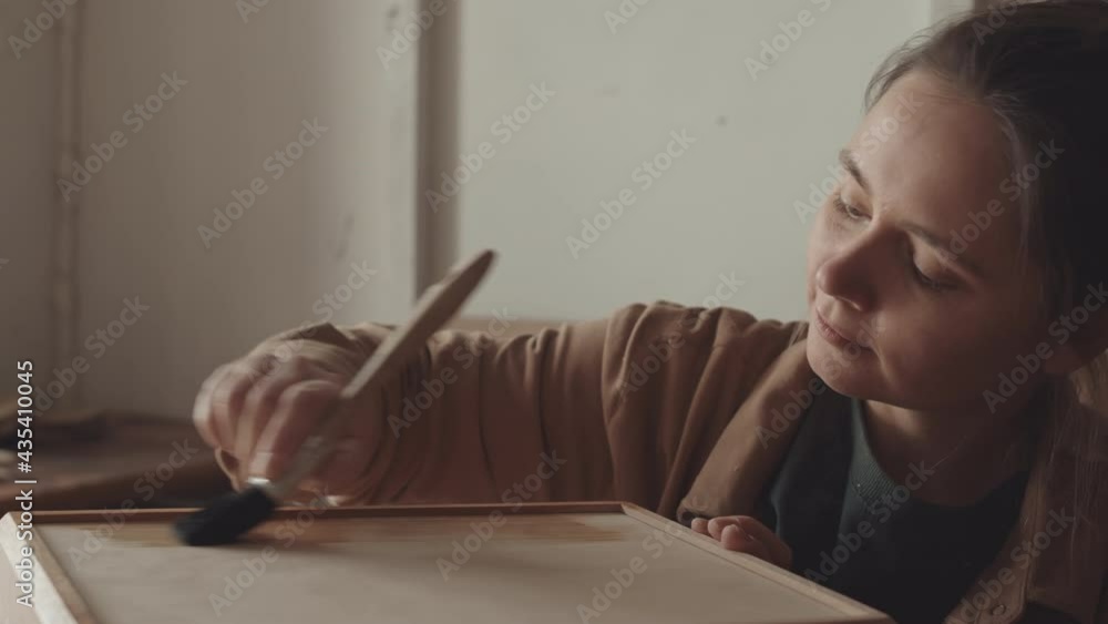 Close up of young Caucasian female carpenter applying wood varnish with brush on handmade box in joinery workshop Close up of young Caucasian female carpenter applying wood varnish with brush on handmade box in joinery workshop