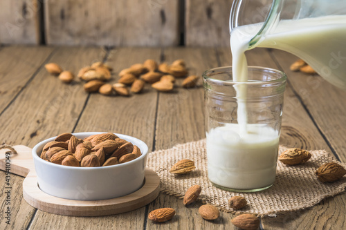Vegan almond milk pouring into a glass, almond kernels and whole almonds on the old rustic wooden table