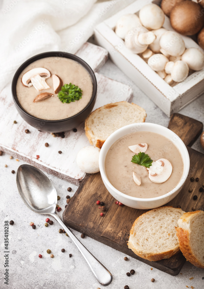 Ceramic bowl plates of creamy chestnut champignon mushroom soup with spoon, pepper and kitchen cloth on white kitchen  background and box of raw mushrooms and fresh bread.