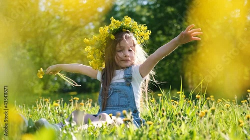 Little cute girl in a flower wreath on her head sits on a sunny meadow with dandelions in her hands