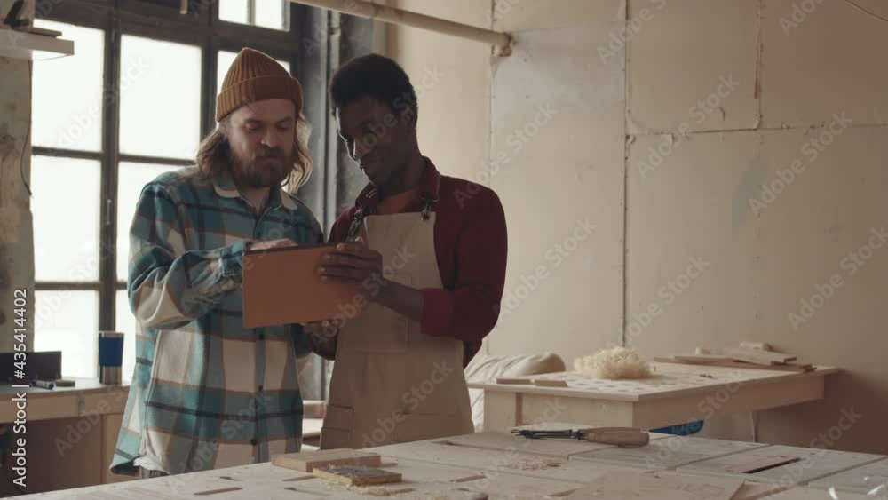 Medium shot of young bearded Caucasian man and African-American man wearing apron standing in carpentry workshop and looking at digital tablet screen Medium shot of young bearded Caucasian man and African-American man wearing apron standing in carpentry workshop and looking at digital tablet screen