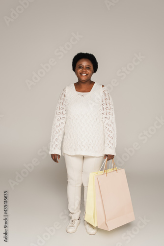 smiling african american middle aged woman in white clothes standing with shopping bags on grey background