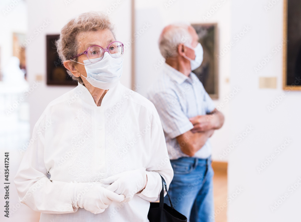 Obraz premium elderly European woman in mask protecting against covid examines paintings on display in hall of art museum