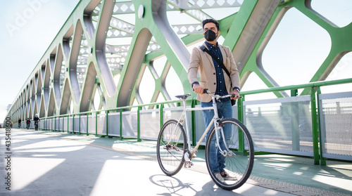 Young business man commuter with bicycle going to work outdoors on bridge in city, coronavirus concept.