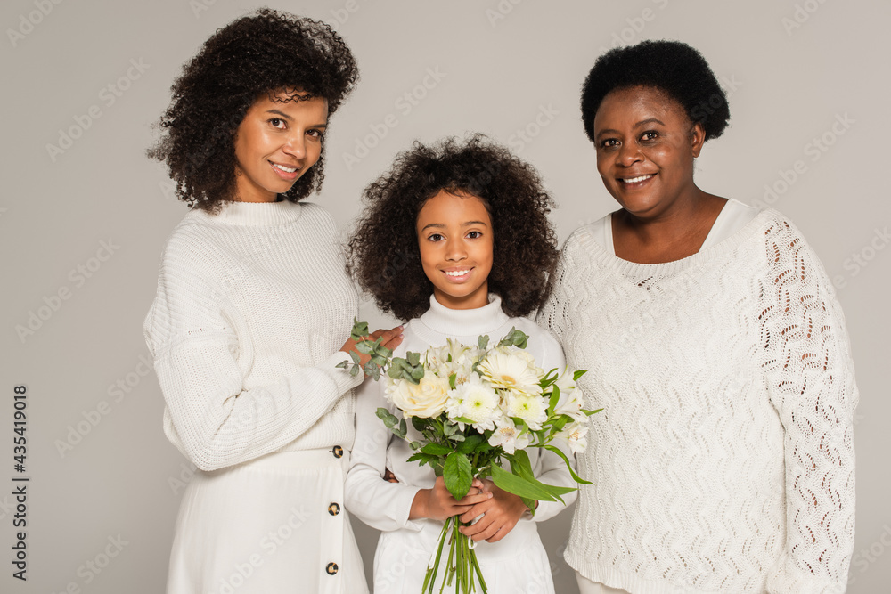 smiling african american mother and grandmother hugging smiling granddaughter with bouquet of flowers isolated on grey