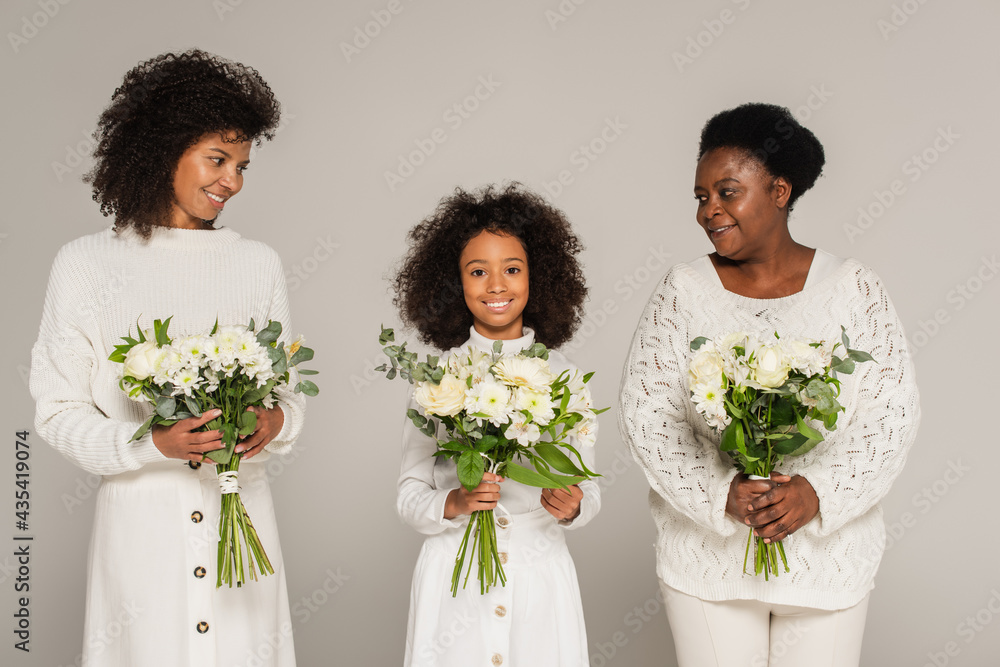 smiling african american mother and grandmother looking at granddaughter and holding bouquets of flowers isolated on grey