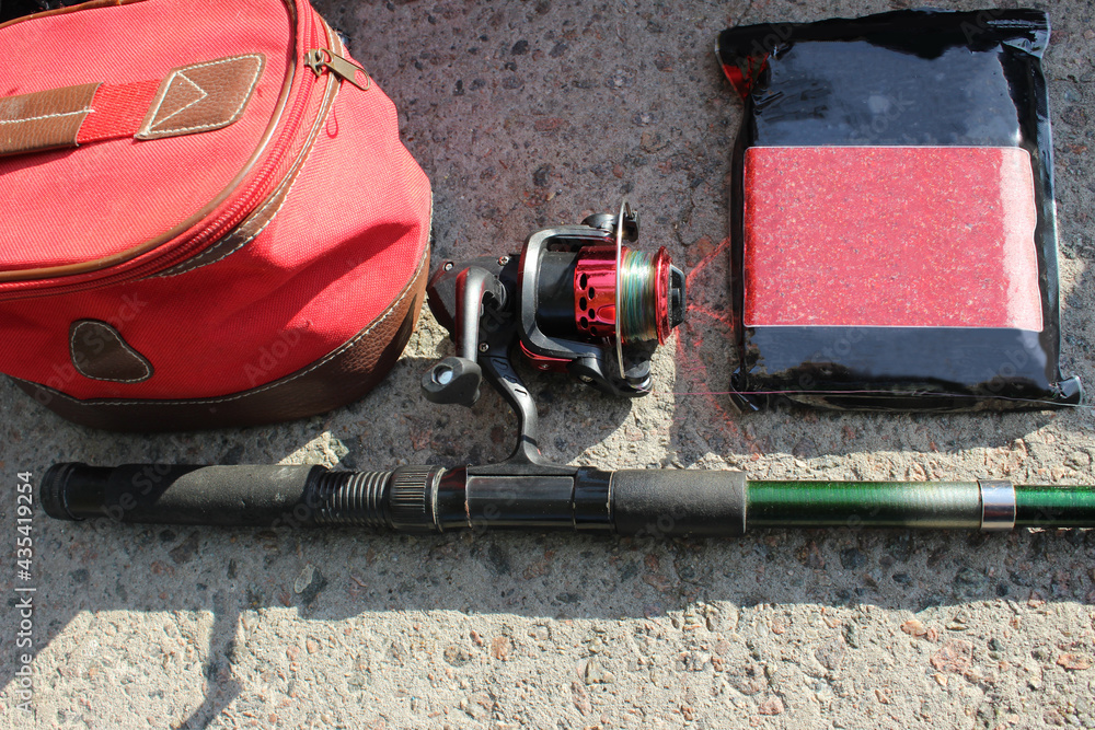 Close-up of a fishing rod with a red reel and colorful line, a fishing bag and fish bait in a rectangular plum-flavored package