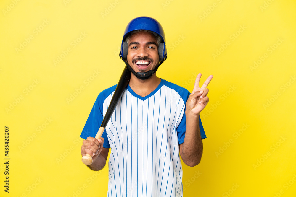 Young Colombian latin man playing baseball isolated on yellow background smiling and showing victory sign Young Colombian latin man playing baseball isolated on yellow background smiling and showing victory sign
