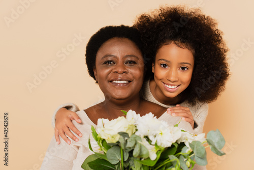 happy african american grandmother and granddaughter with bouquet of flowers isolated on beige