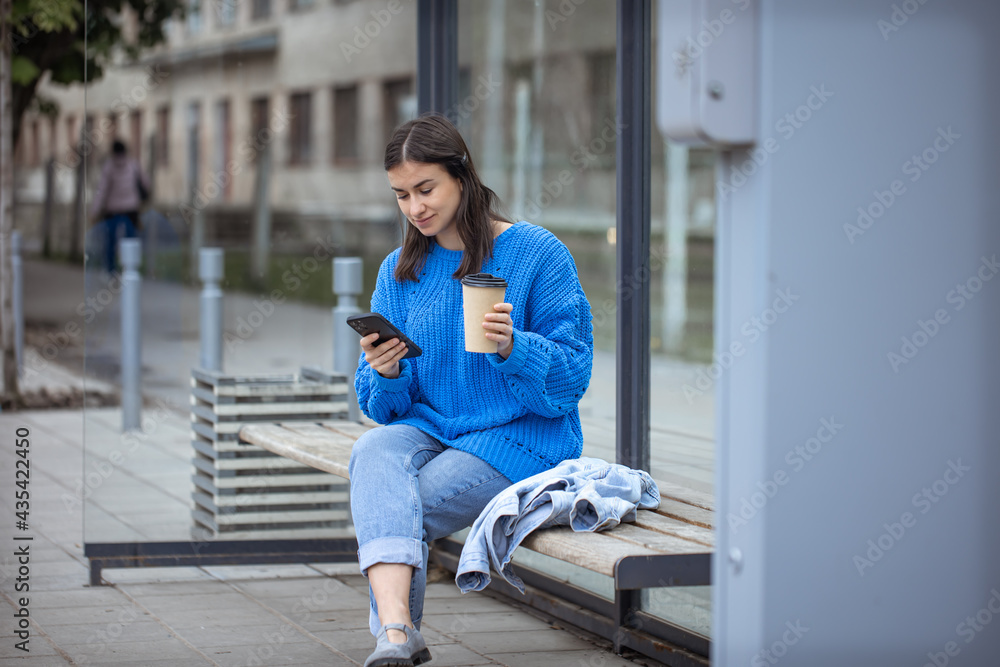 Stylish young girl at the bus stop with coffee in hand, uses the phone.