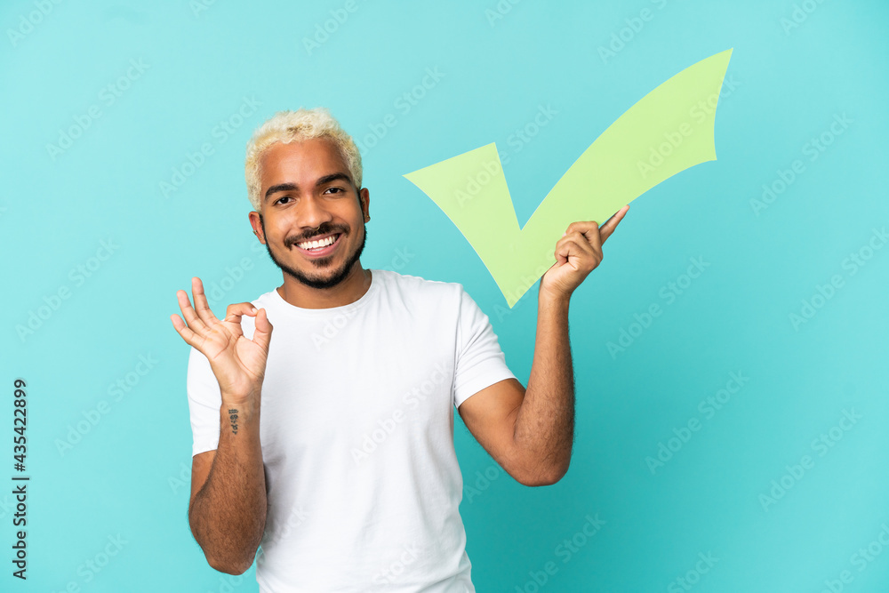 Young Colombian handsome man isolated on blue background holding a check icon and doing OK sign Young Colombian handsome man isolated on blue background holding a check icon and doing OK sign
