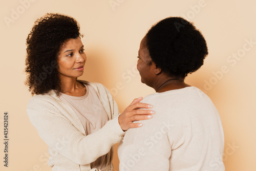 african american adult daughter supporting middle aged mother with hands on shoulders on beige background