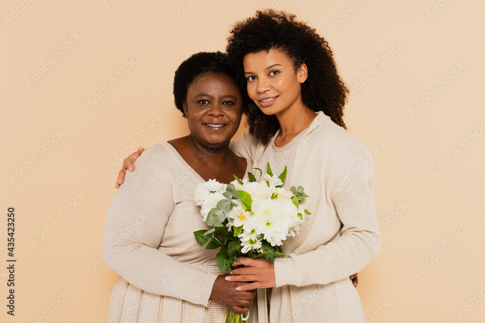 african american adult daughter and middle aged mother standing with bouquet of flowers isolated on beige african american adult daughter and middle aged mother standing with bouquet of flowers isolated on beige