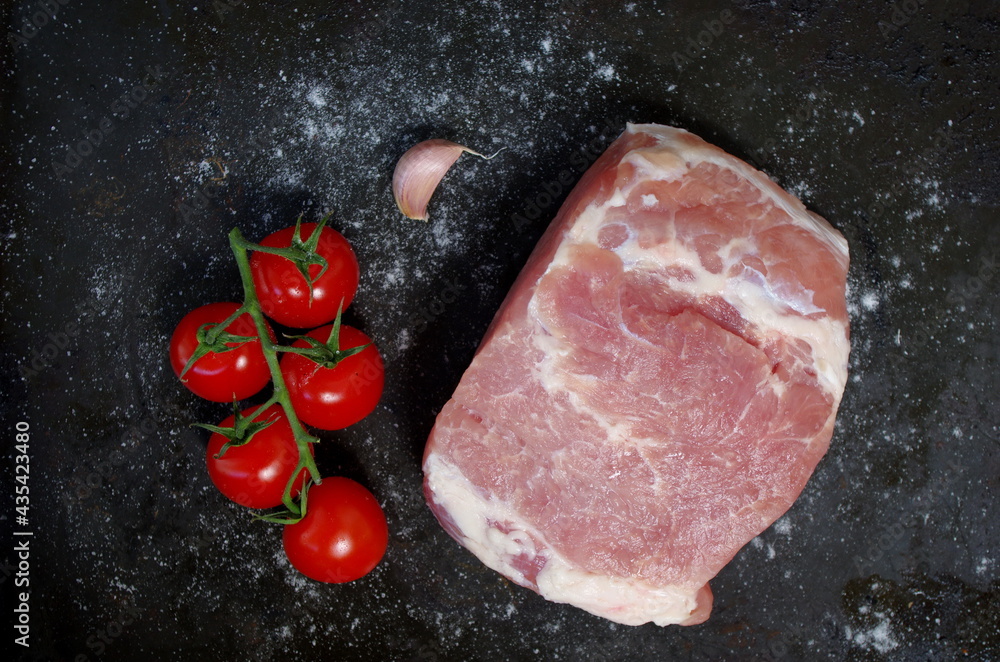 Raw piece of pork with red tomatoes, fresh garlic and salt on black background. Overhead view of Piece of fresh boneless pork, neck part or collar. Big piece of red raw meat on a black background. Raw piece of pork with red tomatoes, fresh garlic and salt on black background. Overhead view of Piece of fresh boneless pork, neck part or collar. Big piece of red raw meat on a black background.