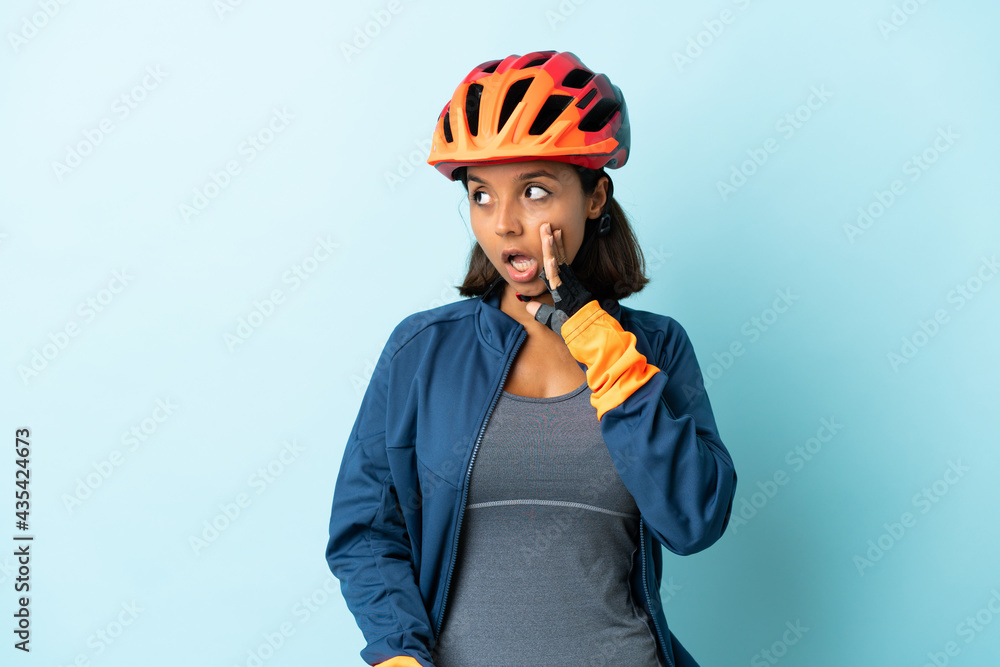 Young cyclist woman isolated on blue background whispering something with surprise gesture while looking to the side - obrazy, fototapety, plakaty 