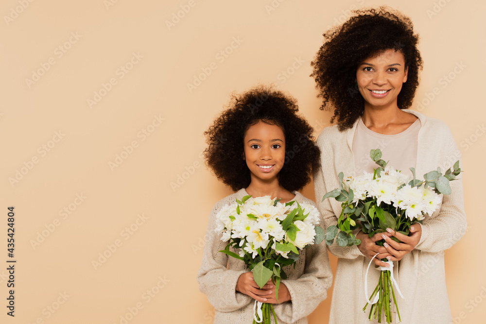 african american preteen daughter and adult mother holding bouquets of daisies isolated on beige