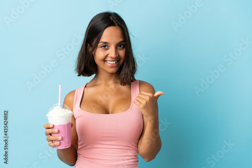 Young woman with strawberry milkshake isolated on blue background pointing to the side to present a product © luismolinero
