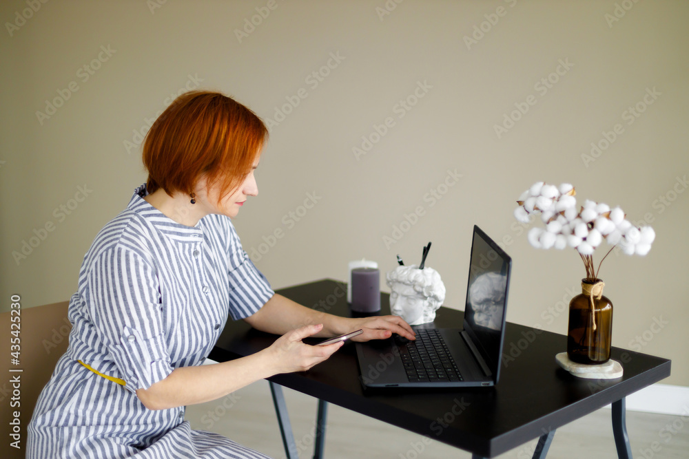 red haired woman in striped dress sits against background of beige wall, works at a laptop. Distant work. Online training. Home office red haired woman in striped dress sits against background of beige wall, works at a laptop. Distant work. Online training. Home office