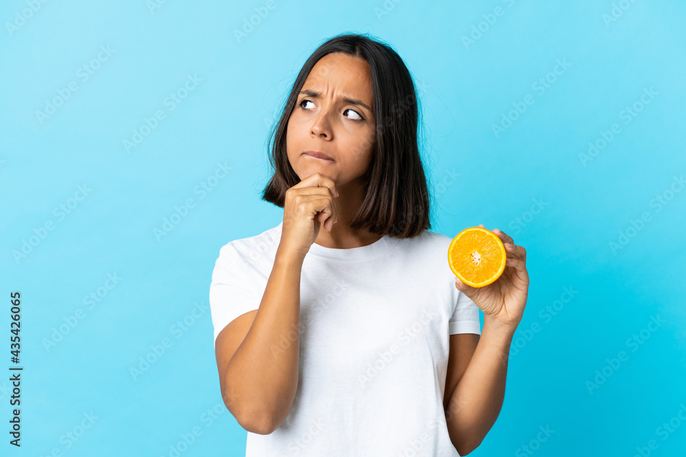 Young asian girl holding an orange isolated on blue background having doubts and thinking Young asian girl holding an orange isolated on blue background having doubts and thinking