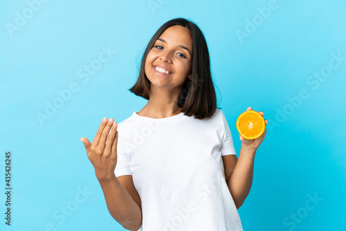 Young asian girl holding an orange isolated on blue background inviting to come with hand. Happy that you came
