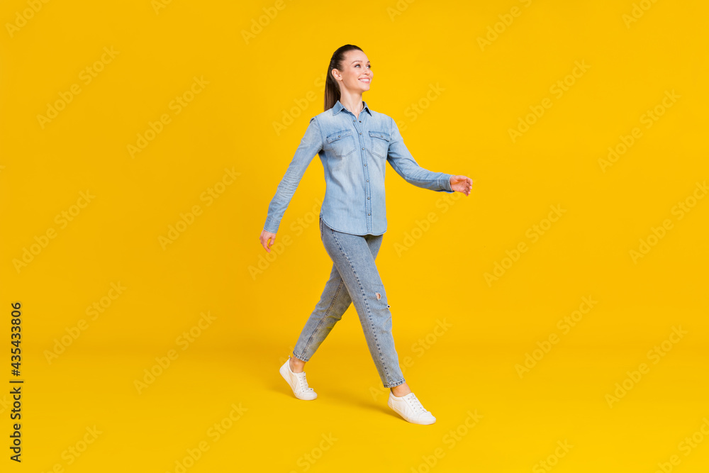 Full length photo of excited sweet young lady dressed jeans shirt walking looking empty space smiling isolated yellow color background