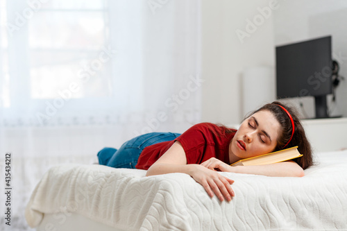 A young Caucasian woman lies on a bed and sleeps with a book under her head. Copy space. White interior of the room on the background. Concept of education and training