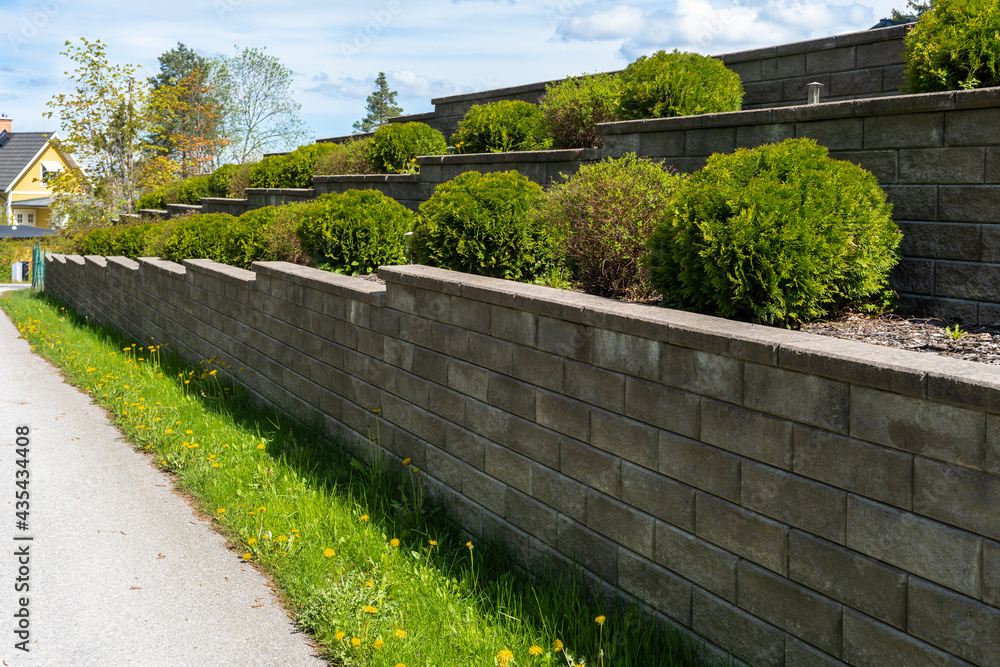 Garden landscape design. Decorative wall made of dark gray stone blocks or concrete cement bricks. Garden decorative trimmed shrubs or bushes are fenced with rocky stones masonry.