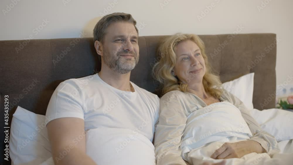 Happy middle-aged couple lying in bed and watching tv, resting together, family Happy middle-aged couple lying in bed and watching tv, resting together, family
