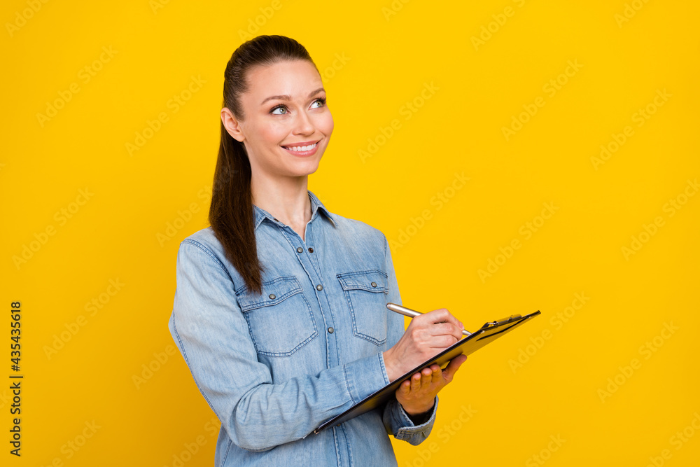 Photo of cute sweet young woman wear denim shirt smiling looking empty space writing clipboard isolated yellow color background Photo of cute sweet young woman wear denim shirt smiling looking empty space writing clipboard isolated yellow color background