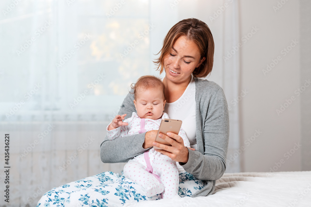 A mother holds a sleeping child in her arms and shows him something on her smartphone. Close-up portrait. Home interior A mother holds a sleeping child in her arms and shows him something on her smartphone. Close-up portrait. Home interior