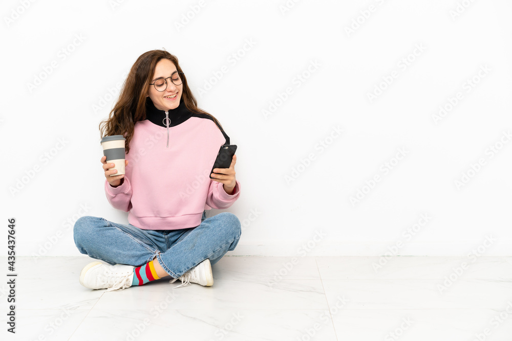 Young caucasian woman sitting on the floor isolated on white background holding coffee to take away and a mobile