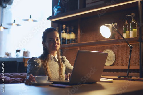 Young beautiful woman sits at a wooden table in a cafe indoors and works on a laptop. Conception of remote work and freelance work.