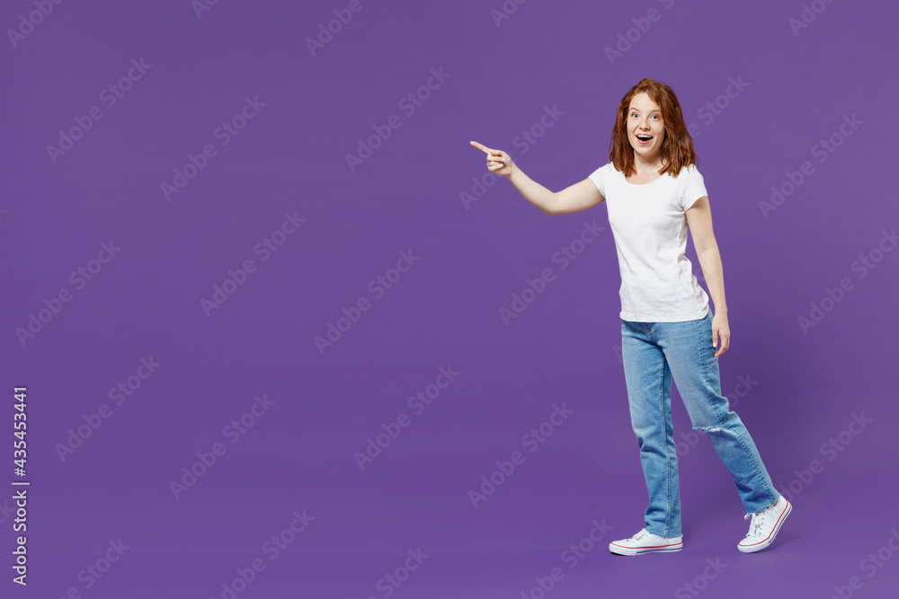 Full length young smiling caucasian woman 20s in white basic casual t-shirt jeans walking going point index finger aside on workspace area mock up isolated on dark violet background studio portrait Full length young smiling caucasian woman 20s in white basic casual t-shirt jeans walking going point index finger aside on workspace area mock up isolated on dark violet background studio portrait