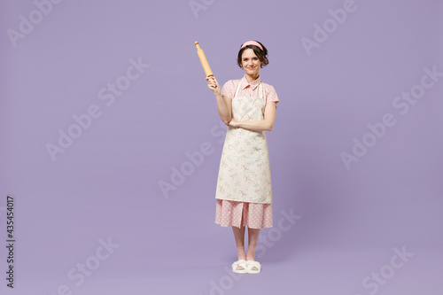 Full length smiling young happy housewife housekeeper chef cook baker woman in pink apron hold rolling pin stand look camera isolated on pastel violet background studio Cooking food process concept