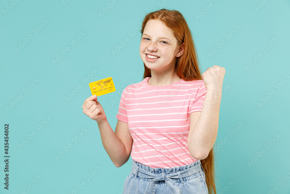 Little redhead kid girl 12-13 years old in pink striped t-shirt holding credit bank card clench fist do winner gesture isolated on pastel blue background studio. Children lifestyle childhood concept Little redhead kid girl 12-13 years old in pink striped t-shirt holding credit bank card clench fist do winner gesture isolated on pastel blue background studio. Children lifestyle childhood concept