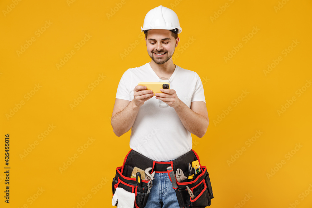 Young employee handyman man in protective helmet hardhat play racing mobile cell phone gadget smartphone isolated on yellow background Instruments accessories renovation apartment room. Repair concept Young employee handyman man in protective helmet hardhat play racing mobile cell phone gadget smartphone isolated on yellow background Instruments accessories renovation apartment room. Repair concept