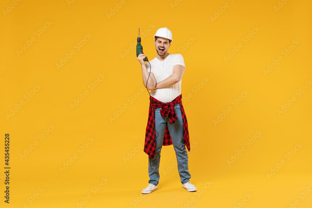 Full length young happy employee handyman man in protective helmet hardhat glasses use electric drill isolated on yellow background Instruments accessory renovation apartment room Repair home concept. Full length young happy employee handyman man in protective helmet hardhat glasses use electric drill isolated on yellow background Instruments accessory renovation apartment room Repair home concept.