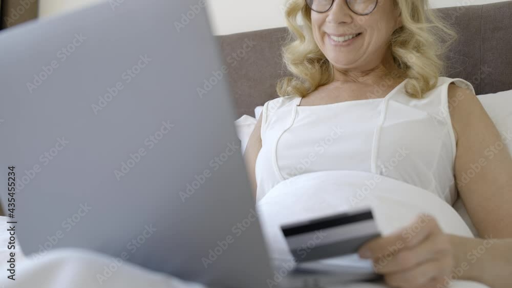 Happy middle-aged lady shopping online in bed, entering card number on laptop Happy middle-aged lady shopping online in bed, entering card number on laptop