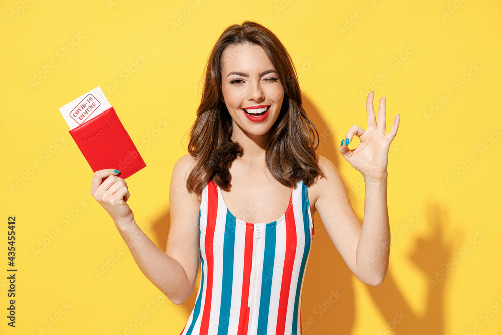 Happy fun young woman slim body wear red blue swimsuit hold passport covid-19 immune vaccination isolated on vivid yellow color background studio Summer hotel pool sea rest sun tan air flight concept. Happy fun young woman slim body wear red blue swimsuit hold passport covid-19 immune vaccination isolated on vivid yellow color background studio Summer hotel pool sea rest sun tan air flight concept.