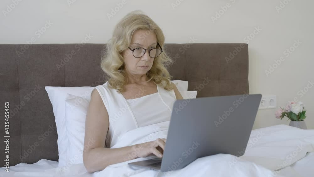 Mature female freelancer in eyeglasses working on laptop in bed, remote work