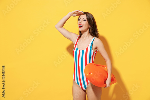 Happy young sexy lifeguard woman slim body wear striped red blue one-piece swimsuit hold aid lifebuoy isolated on vivid yellow color wall background studio. Summer hotel pool sea rest sun tan concept.