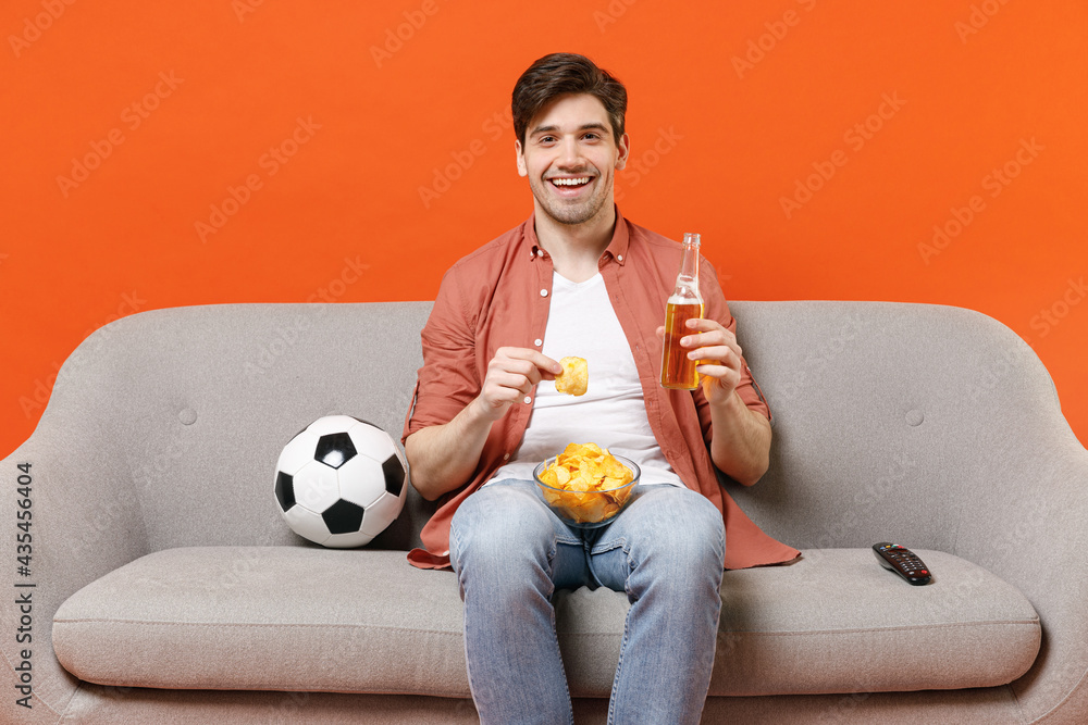 Young smiling happy man football fan in shirt support team with soccer ball sit home sofa watch tv live stream eat chips drink beer isolated on orange background studio People leisure sport concept