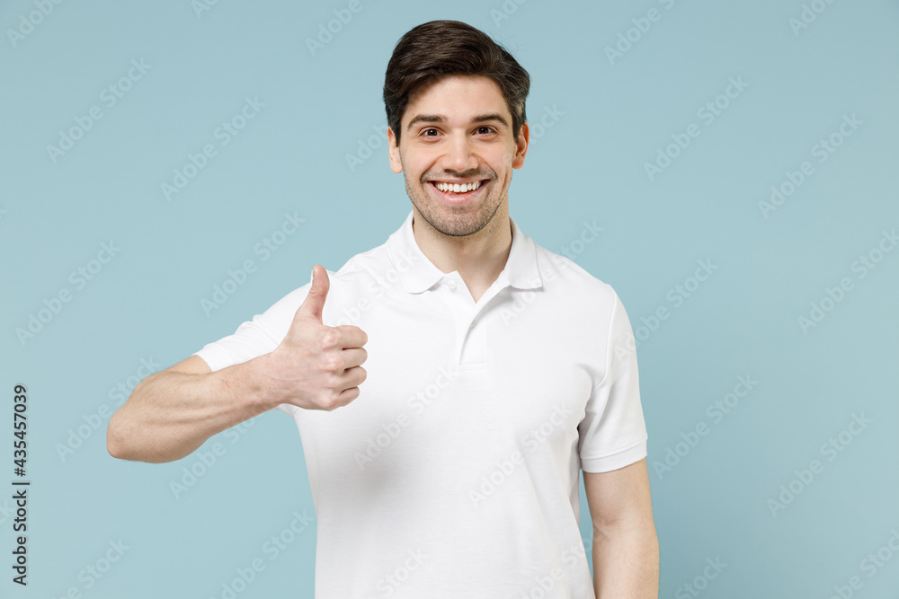 Young fun happy smiling unshaven cheerful caucasian man 20s wear white casual basic t-shirt show thumb up gesture isolated on pastel blue color background studio portrait. People lifestyle concept Young fun happy smiling unshaven cheerful caucasian man 20s wear white casual basic t-shirt show thumb up gesture isolated on pastel blue color background studio portrait. People lifestyle concept