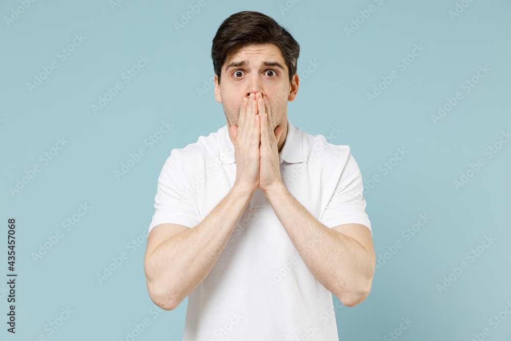 Young shocked amazed scared confused unshaven caucasian man 20s in white casual basic t-shirt cover mouth with hand isolated on pastel blue color background studio portrait. People lifestyle concept