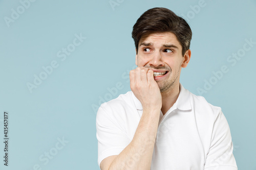 Young confused puzzled thoughtful pensive unshaven caucasian man 20s in white casual basic t-shirt look aside biting nails isolated on pastel blue background studio portrait. People lifestyle concept.